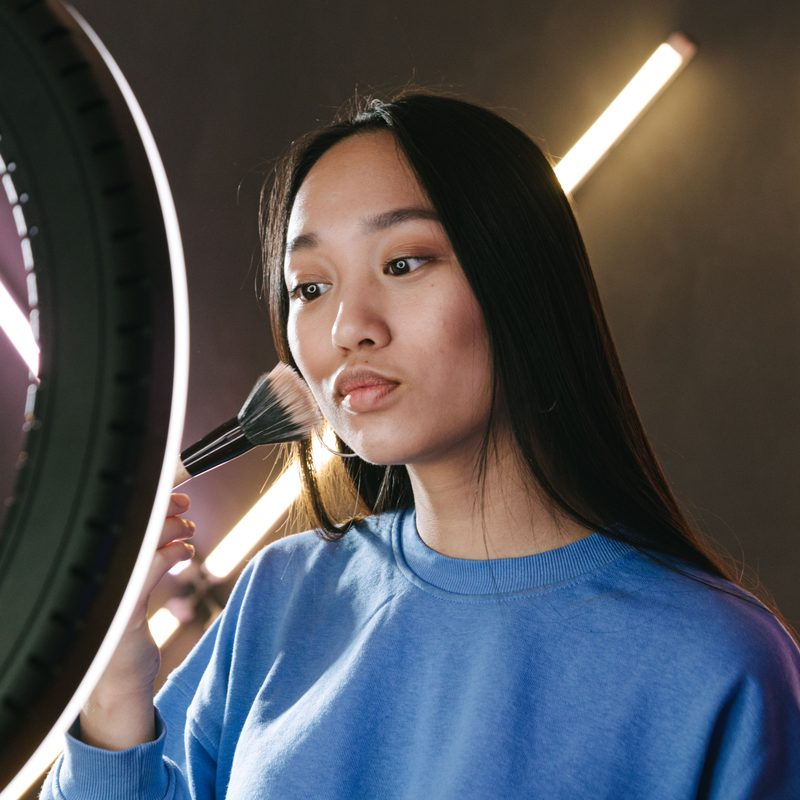Creator in a blue sweatshirt applying makeup in front of a ring light, preparing content for X without getting shadowbanned
